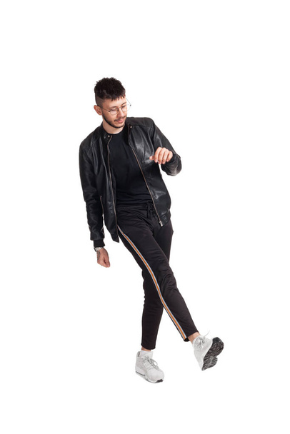 Full-length foto van een moderne artiest in bril, zwart leren jasje, t-shirt, sportbroek en lichte sneakers die in de studio rondlopen. Indoor foto van een sterke collega dansen en naar beneden kijken - Foto, afbeelding