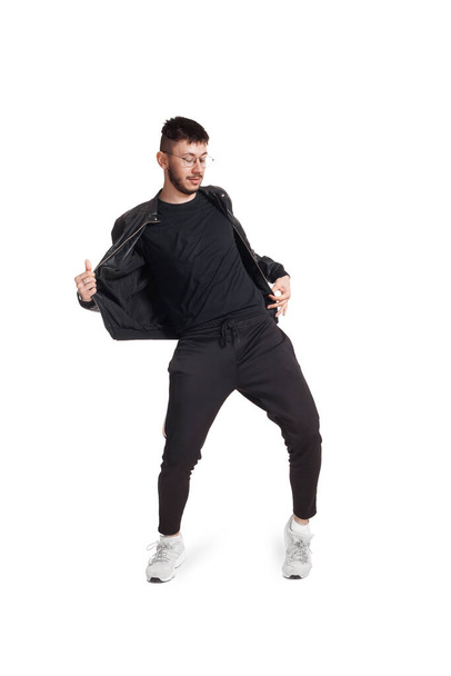 Full-length photo of a modern performer in glasses, black leather jacket, t-shirt, sports pants and light sneakers fooling around in studio. Indoor photo of an athletic person dancing and looking down - Photo, Image