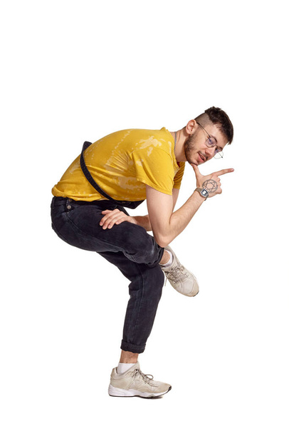 Retrato de cuerpo entero de un guapo hombre con gafas, mono negro, camiseta amarilla y zapatillas grises jugando en el estudio. Foto interior de un hombre haciendo elementos de baile aislados en blanco - Foto, imagen
