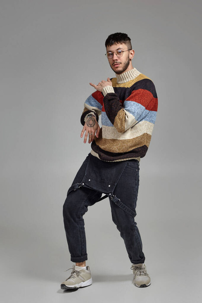 Full-length portrait of a good-looking man in glasses, black jumpsuit, multi-colored sweater and gray sneakers fooling around in studio. Indoor photo of a man dancing on a gray background. Music and - Photo, Image