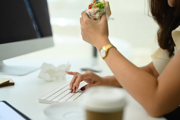 Colpo ritagliato di impiegato donna seduto alla scrivania e mangiare panino per pranzo. - Foto, immagini