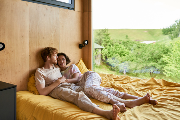 redhead man lying on bed with cheerful asian girlfriend at looking at window with natural view - Photo, Image