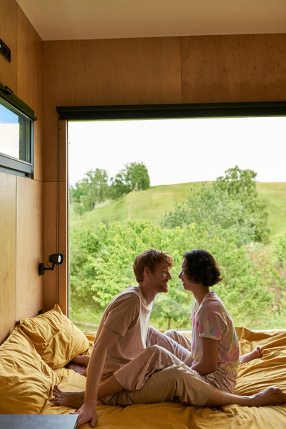 happy diverse couple looking at each other and sitting on bed in country house with forest view - 写真・画像