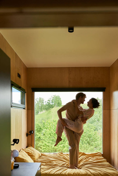 beztroska wielokulturowa para tańcząca razem na łóżku w wiejskim domu, okno z widokiem na las - Zdjęcie, obraz