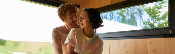 joyful redhead man hugging asian girlfriend next to window with forest view in country house, banner - Foto, imagen