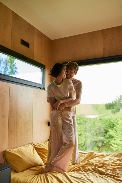 happy man hugging asian girlfriend standing on bed near window with forest view in country house - Fotografie, Obrázek