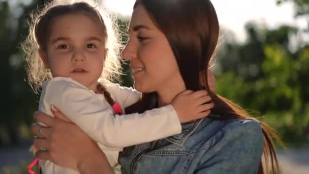 Close-up portrait of happy young woman kissing little girl talking with kid standing in sunrays. Confident beautiful Caucasian mother enjoying leisure with daughter at sunset in summer spring park - Filmagem, Vídeo