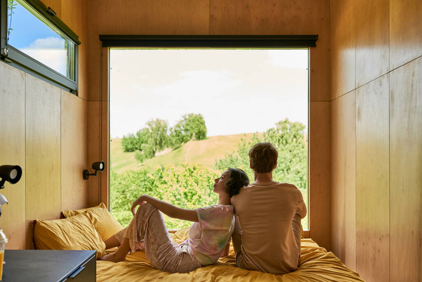 Asyalı kadın yatakta oturuyor ve erkek arkadaşının omzuna yaslanıyor, kır evinde birlikte rahatlıyor. - Fotoğraf, Görsel