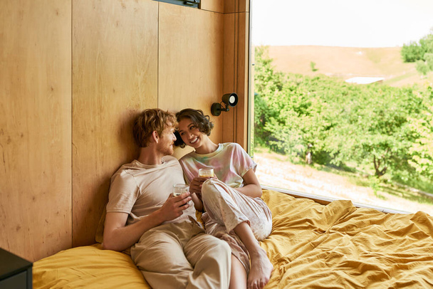 onnellinen rotujenvälinen pari istuu sängyllä ja tilalla kuppeja aamulla kahvia Country House - Valokuva, kuva