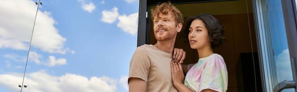 casal interracial sonhador em pé na porta da frente da casa de vidro moderno e olhando para longe, banner - Foto, Imagem