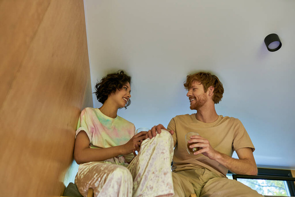 szczęśliwy Azji kobieta i ruda mężczyzna gospodarstwa filiżanki kawy i siedzi razem w przytulne łóżko piętrowe - Zdjęcie, obraz