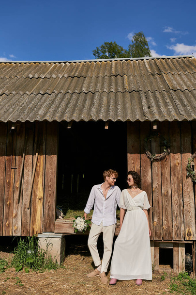 concepto de boda rústico interracial recién casados en gafas de sol y vestido de novia cerca de granero de madera - Foto, imagen