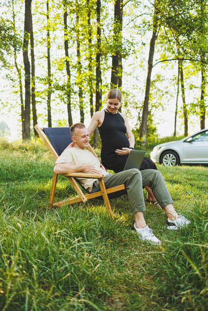 A happy man and his pregnant wife are working on a laptop while sitting in nature. A young couple is relaxing in nature and watching a movie on a laptop. - Valokuva, kuva