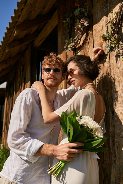 azjatycka kobieta w sukni ślubnej przytula się z panem młodym w okularach przeciwsłonecznych w pobliżu drewnianej stodoły, rustykalny ślub - Zdjęcie, obraz
