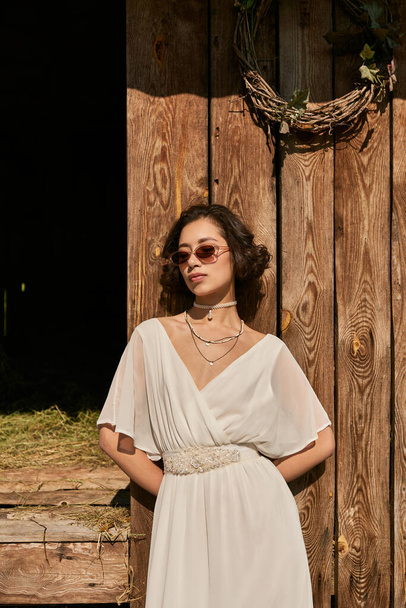 giovane sposa asiatica in abito da sposa bianco e occhiali da sole in piedi vicino fienile di legno in campagna - Foto, immagini