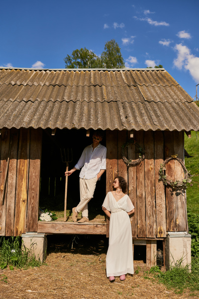 rustic wedding concept, interracial newlyweds posing near wooden barn, couple in wedding gown - Фото, зображення