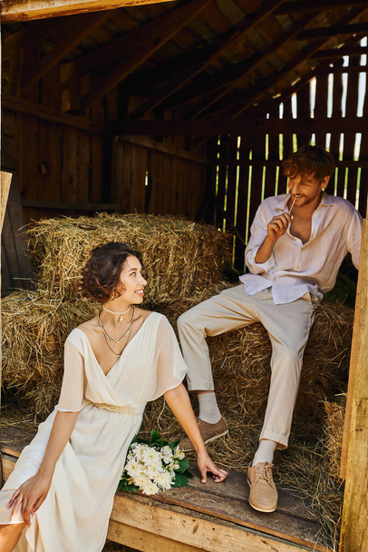 happy asian bride in white dress holding bouquet and sitting on hay near groom in barn, rural - Foto, Imagem