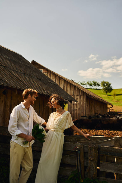 happy asian bride in white dress standing near groom in sunglasses near stable in countryside - Foto, afbeelding