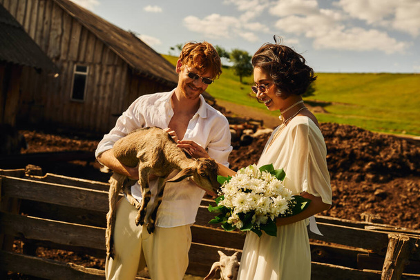 happy multiethnic newlyweds cuddling cute baby goat, asian woman in wedding dress and sunglasses - Photo, Image