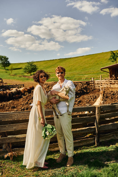 сільське весілля в стилі бохо, щасливий наречений тримає дитину козла біля азіатської нареченої у весільній сукні - Фото, зображення