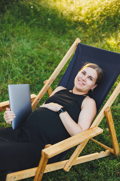 Una linda mujer embarazada está sentada en un parque y trabajando remotamente en un portátil. Trabajo en línea sobre licencia de maternidad. Mujer embarazada moderna - Foto, Imagen
