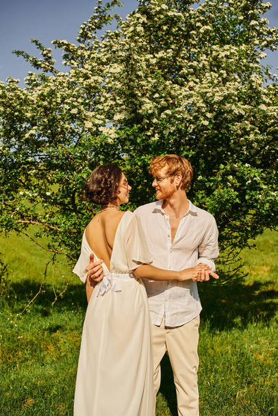 happy newlyweds in countryside, asian bride in white dress and redhead groom hugging near big tree - Фото, изображение