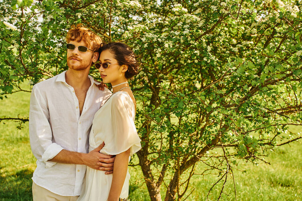 молодые многонациональные молодожены в солнцезащитных очках и свадебных платьях, обнимающиеся в зеленом саду, в стиле бохо - Фото, изображение