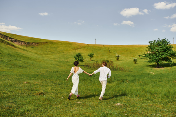 young couple of newlyweds running in green field, bride and groom in wedding gown holding hands - Photo, Image