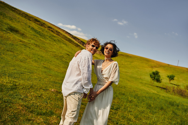 mariage rural à la campagne, jeunes mariés multiethniques en robe de mariée regardant la caméra dans le champ vert - Photo, image