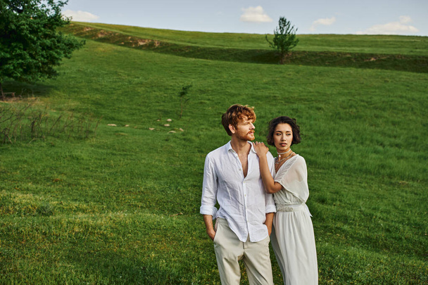 just married interracial couple standing together in green field, scenic and tranquil landscape - Foto, Imagen