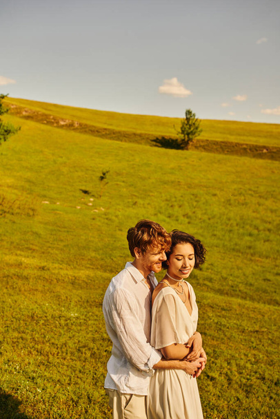 just married couple, happy man hugging asian bride in green field, scenic and tranquil landscape - Foto, imagen