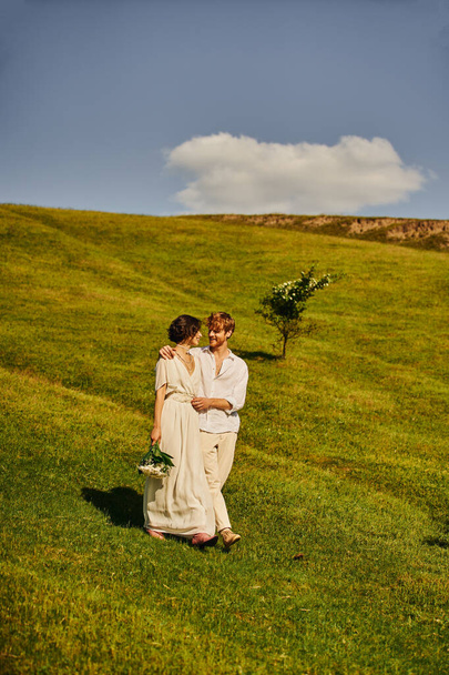 happy interracial newlyweds in boho style wedding attire walking in green field, scenic landscape - Photo, Image