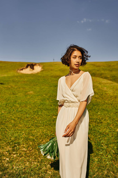 morena asiática mulher no boho estilo casamento vestido segurando flores e olhando para longe no campo verde - Foto, Imagem
