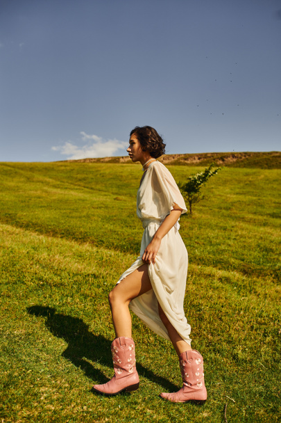 πλαϊνή άποψη της νεαρής Ασιάτισσας με νυφικό φόρεμα και καουμπόικες μπότες περπάτημα στο λιβάδι στην ύπαιθρο - Φωτογραφία, εικόνα
