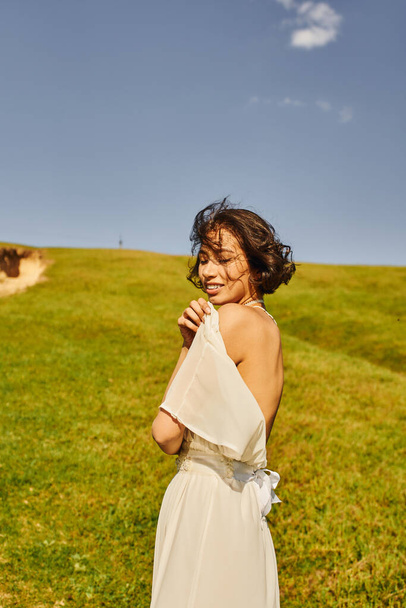 sensual asiático novia en boho estilo vestido sonriendo en verde prado bajo azul cielo, serenidad y alegría - Foto, imagen