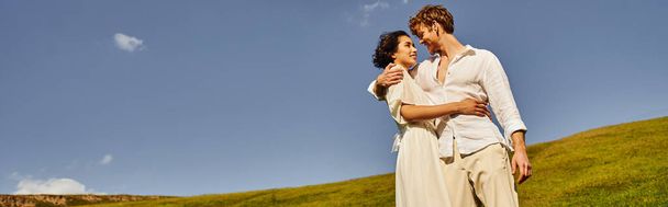 happy multiethnic newlyweds hugging on green meadow under blue sky, wedding in rural setting, banner - Foto, immagini
