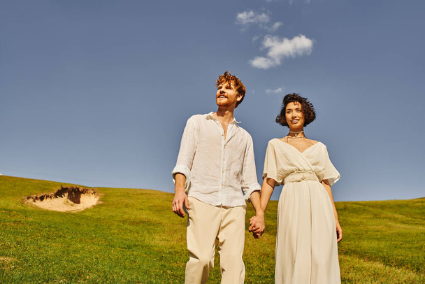 jeunes, insouciants interracial jeunes mariés tenant la main dans le champ vert sous le ciel bleu, paysage pittoresque - Photo, image