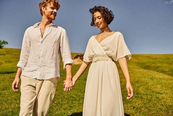 szczęśliwy moment międzyrasowych nowożeńców w stroju boho trzymających się za ręce na polu, wiejskie wesele - Zdjęcie, obraz