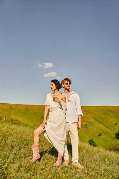 asian woman in wedding dress and cowboy boots near redhead groom in sunglasses in countryside - Photo, Image