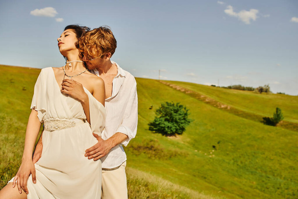 redhead man in sunglasses embracing elegant asian bride on green meadow, tranquil rustic wedding - Photo, Image