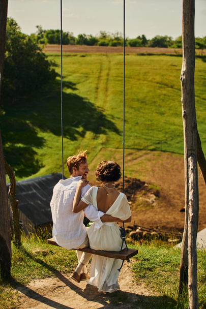 excited redhead groom embracing bride in white dress while having fun on swing in rural setting - Foto, Bild