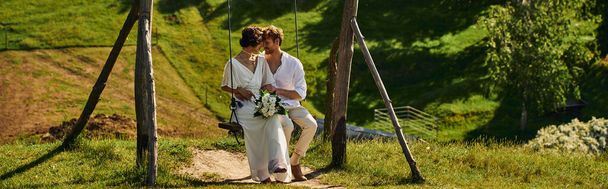 joyful interracial newlyweds in boho style attire on swing in picturesque rural setting, banner - Фото, изображение