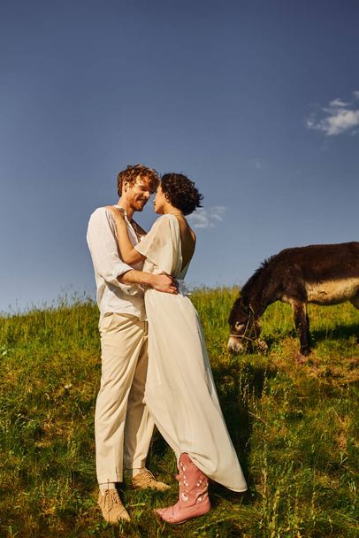 happy redhead man embracing young asian bride near donkey grazing on green meadow, rustic wedding - Photo, Image