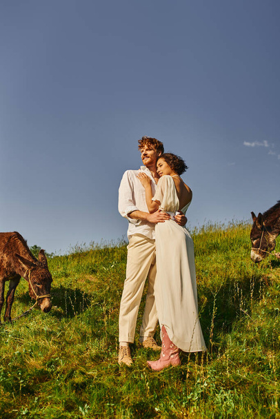 joyful interracial newlyweds embracing and looking away near donkeys grazing on rustic farmland - Photo, Image
