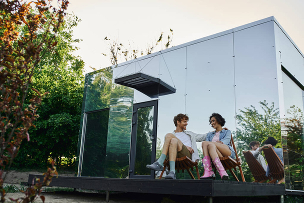 happy multiethnic couple sitting on chairs and looking at each other near modern glass house - Photo, Image