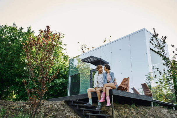 ξέγνοιαστο διαφυλετικό ζευγάρι κάθεται στη βεράντα του σύγχρονου σπιτιού γρασίδι και χαμογελώντας ο ένας στον άλλο - Φωτογραφία, εικόνα