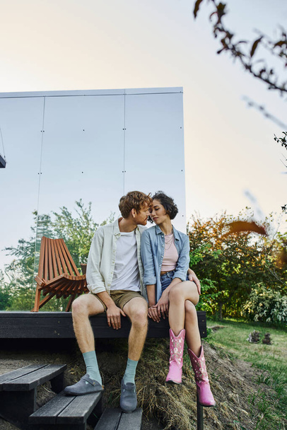 многонациональная пара в стильной повседневной одежде целуется на крыльце стеклянного дома в сельской местности - Фото, изображение