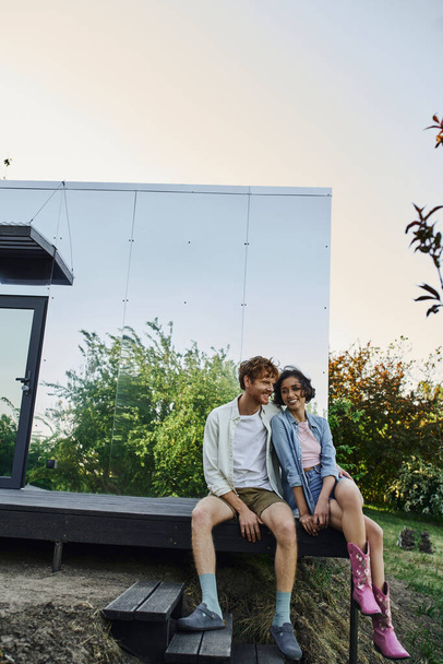 joyful multiethnic couple sitting on porch of modern glass house during weekend in countryside - Photo, Image