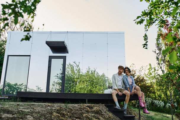 cheerful interracial couple sitting on porch of modern glass house in countryside setting - Photo, Image