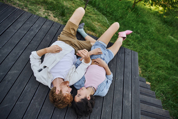 carefree multiethnic couple in stylish casual attire lying down on wooden porch outdoors, top view - Photo, Image